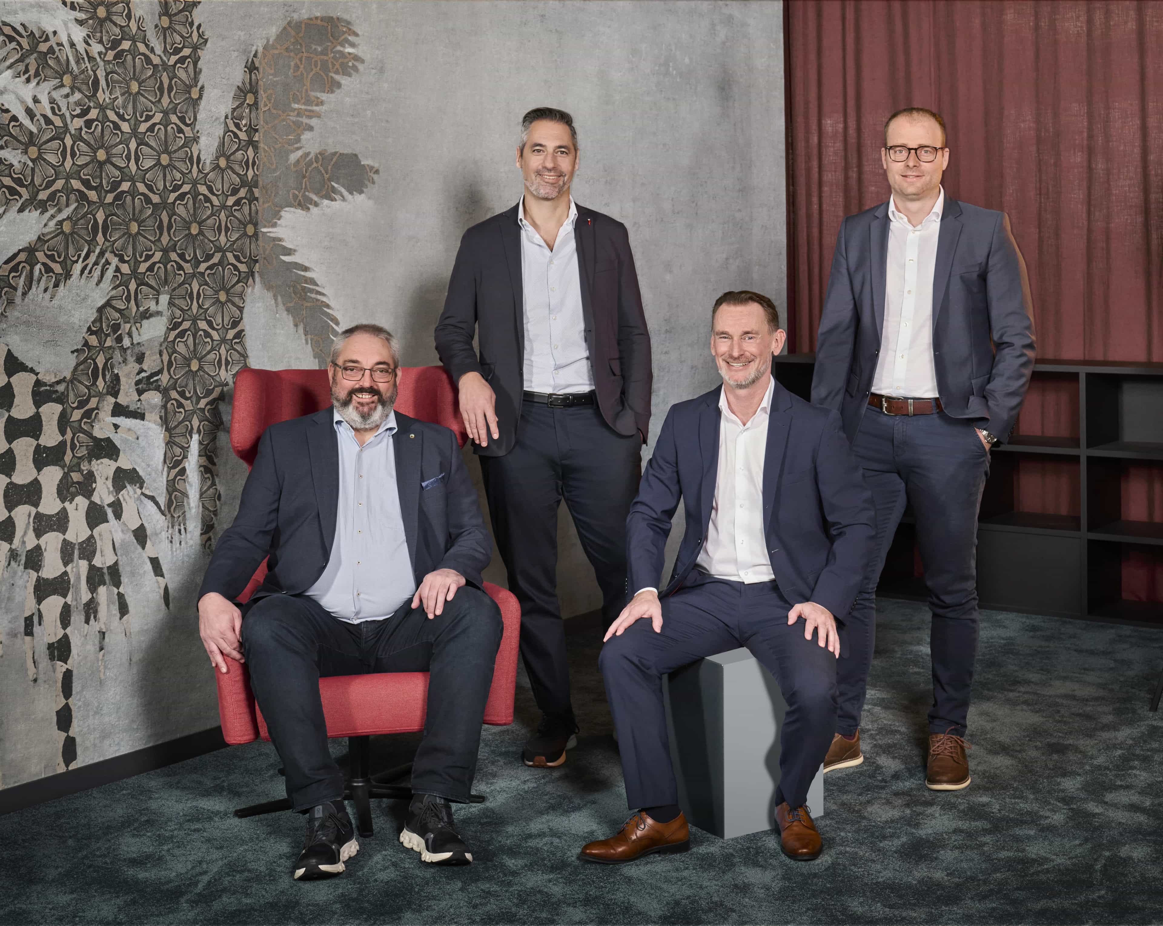 Vier Männer in Anzügen posieren vor einer gemusterten Wand. Zwei sitzen, zwei stehen. Elegante Business-Umgebung.
